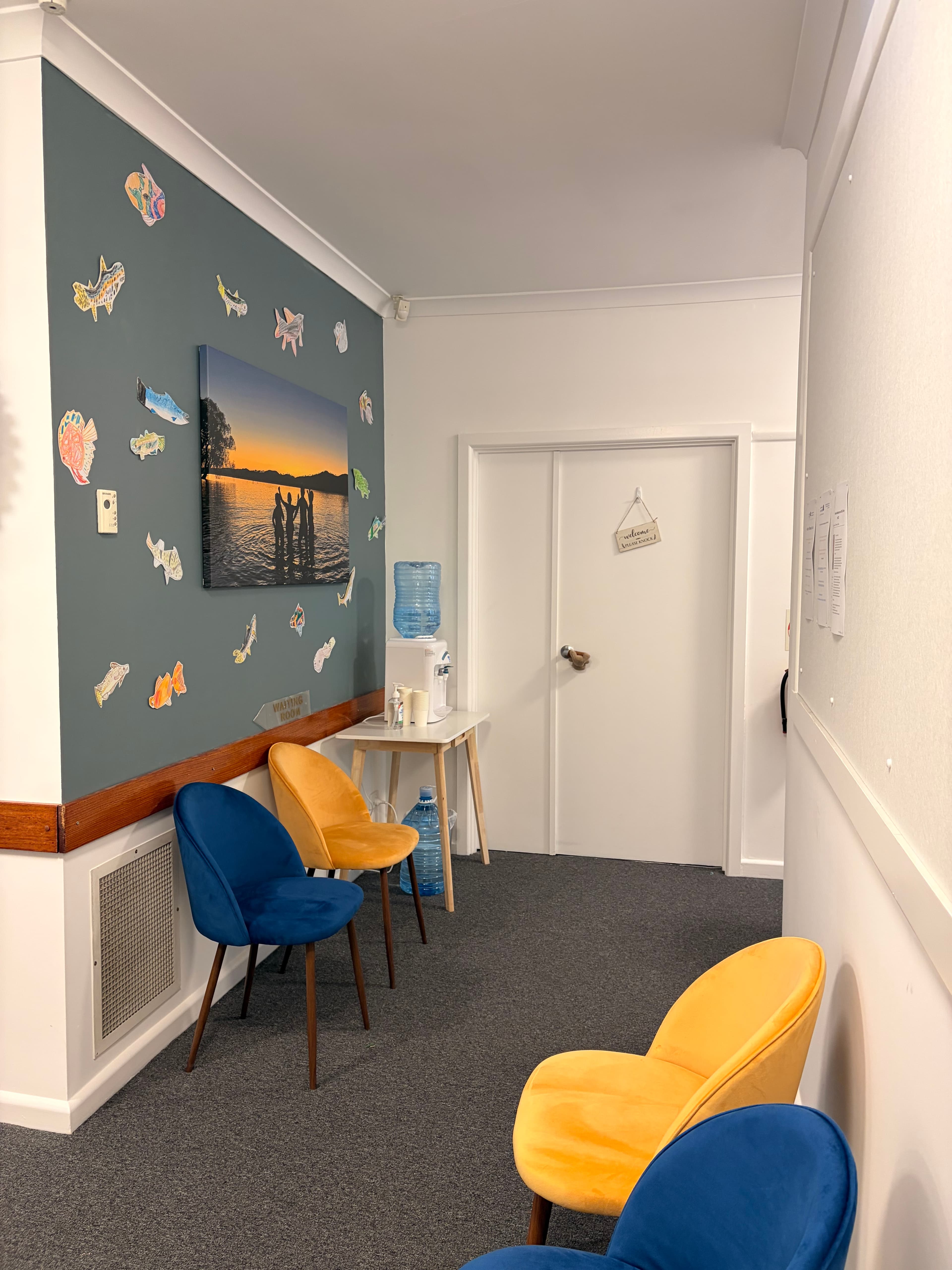 The waiting room at Brain Helpers — comfortable blue and yellow armchairs, a wall of hand-drawn fish around a sunset canvas.